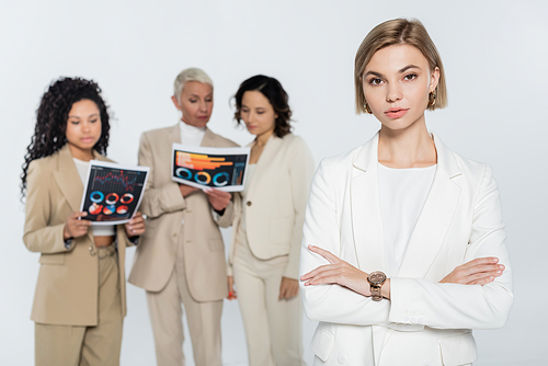 Blonde businesswoman looking at camera near blurred interracial colleagues with papers isolated on grey