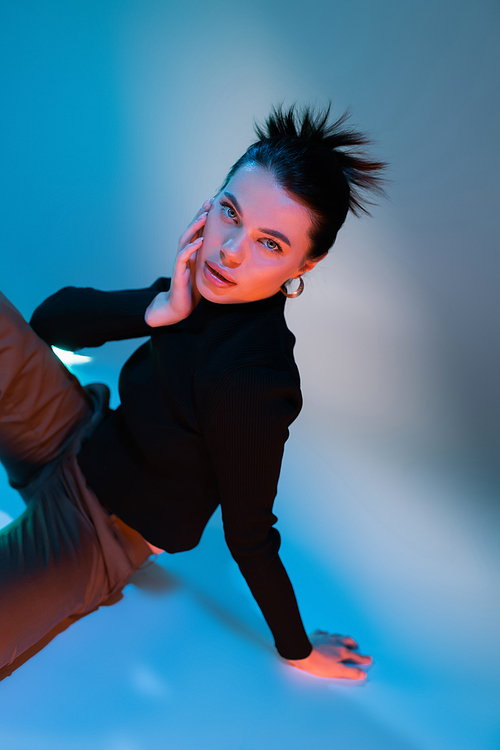 sensual and stylish woman in black sweater touching face and looking at camera on blue background