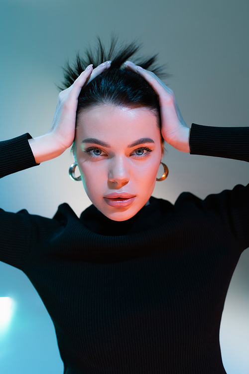 portrait of brunette woman in black sweater and with natural makeup touching hair on grey background