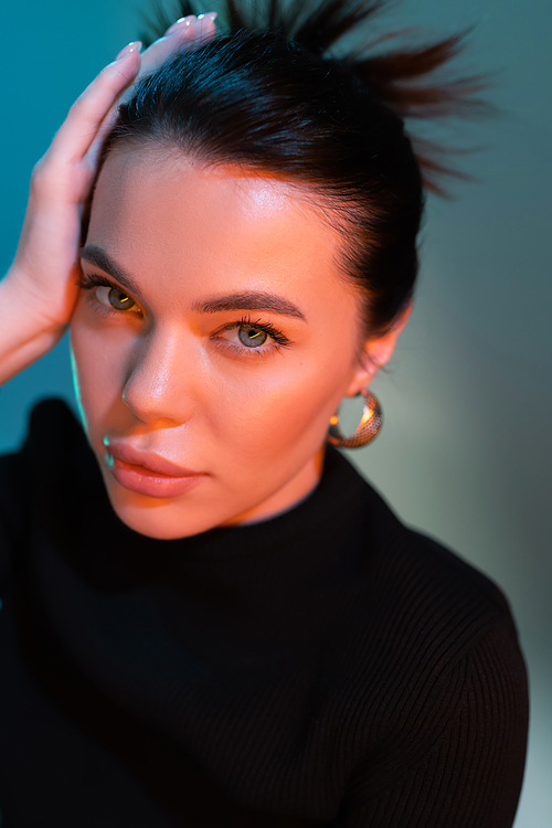 portrait of brunette woman with natural makeup touching hair and looking at camera on grey and blue background