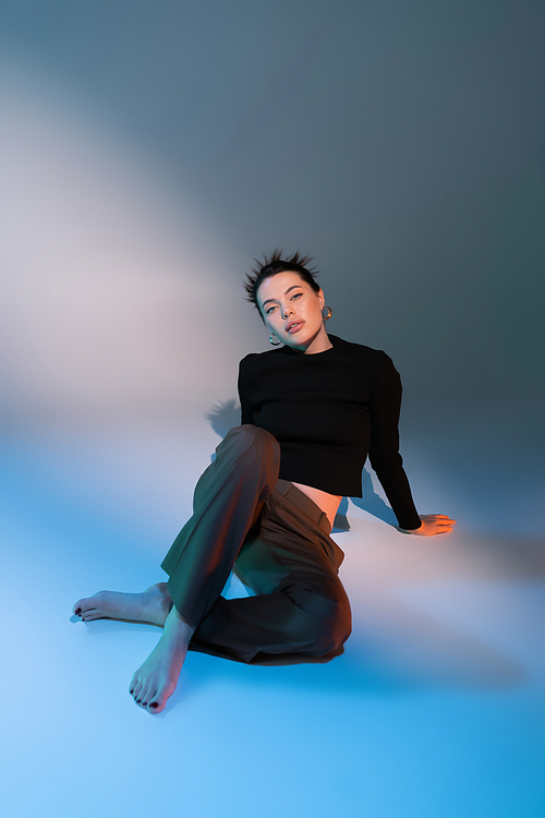 full length of sensual barefoot woman in brown pants and black sweater sitting on gray and blue background