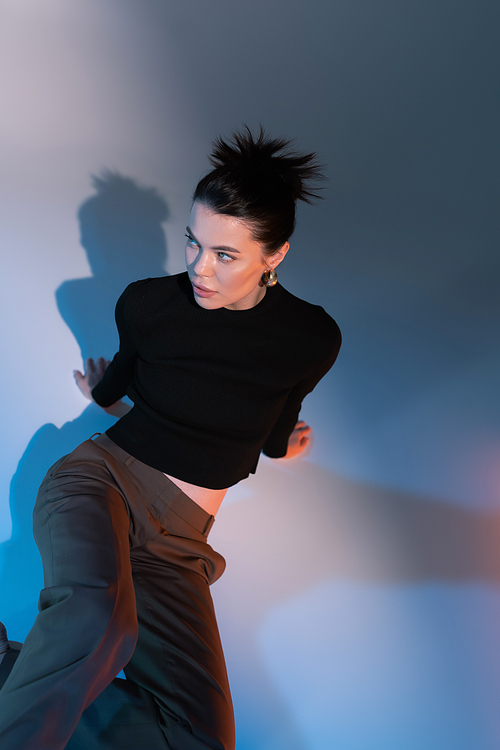 top view of brunette woman in black sweater and brown pants sitting and looking away on grey background