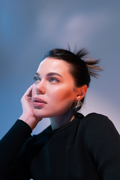 portrait of brunette woman with natural makeup holding hand near face and looking away on blue background