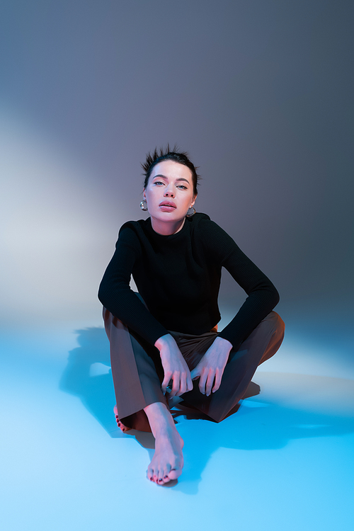 full length of stylish barefoot woman in black sweater looking at camera while sitting on grey and blue background