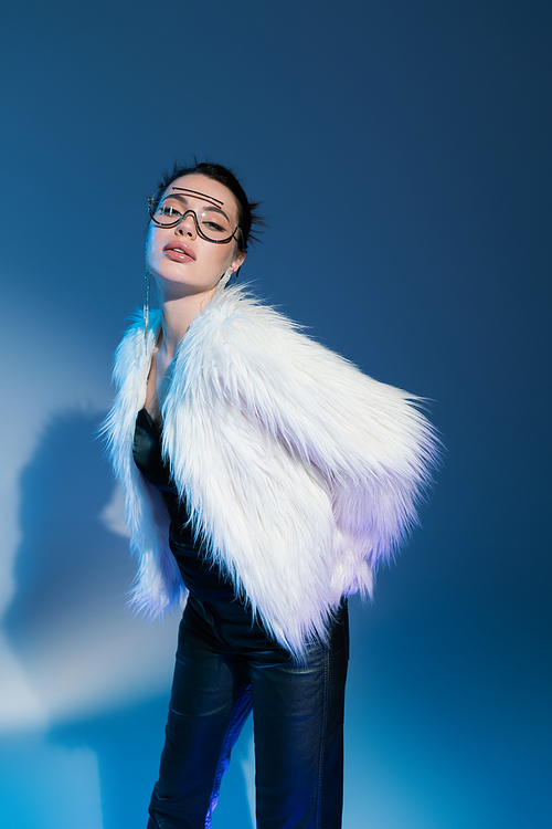 young brunette woman in white faux fur jacket and transparent sunglasses looking at camera while posing on blue background