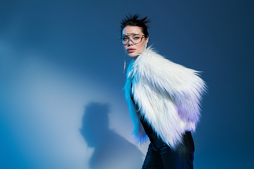 young woman in transparent eyeglasses and fluffy faux fur jacket looking at camera on blue background