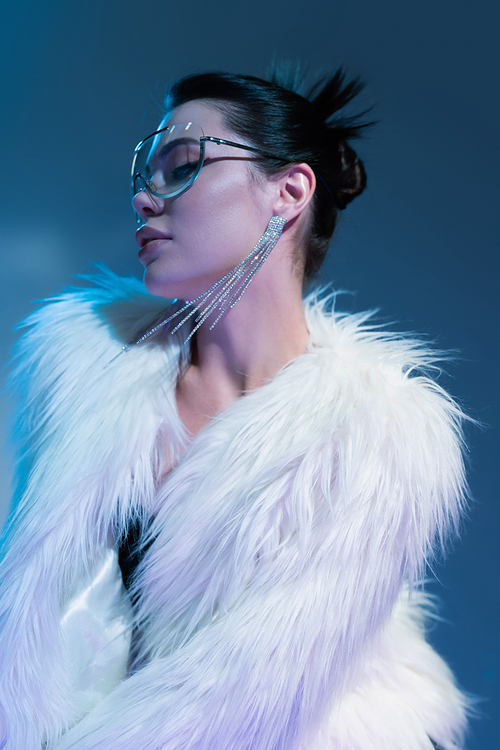 brunette woman in transparent sunglasses and white faux fur jacket on blue background