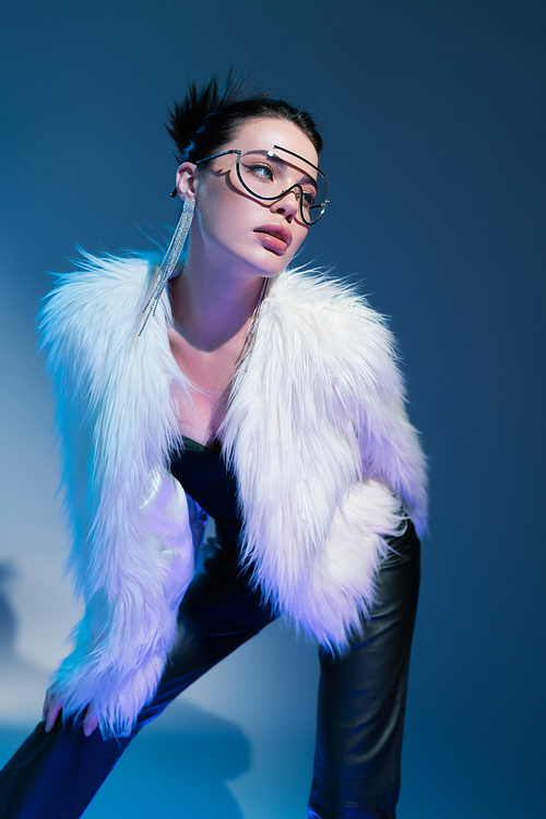 young model in transparent eyeglasses and white faux fur jacket looking away on blue background