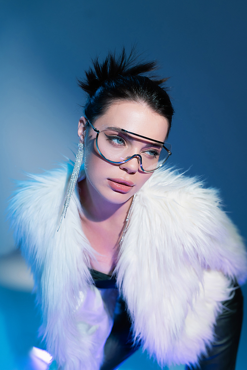 portrait of charming woman in white faux fur jacket and transparent sunglasses looking away on blue background