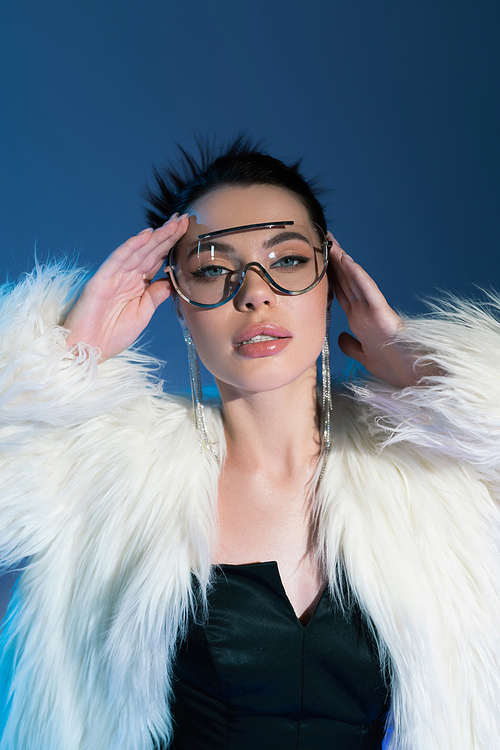 fashionable woman in fluffy faux fur jacket and transparent sunglasses holding hands near face on blue background