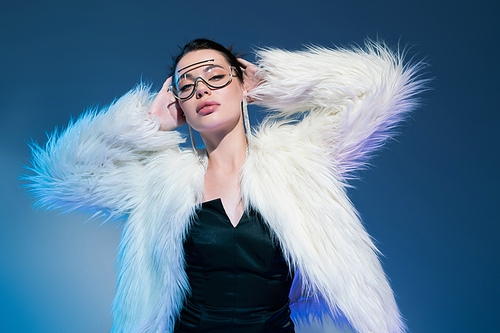 pretty woman in black corset and white faux fur jacket posing with hands behind head on blue background