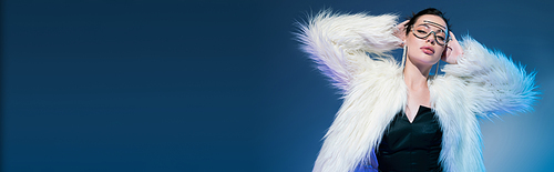 brunette woman in transparent sunglasses and white furry jacket standing with hands behind head on blue background, banner