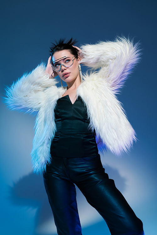 model in black corset and trendy faux fur jacket posing with hands behind head on blue background