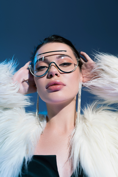 portrait of glamour woman in white fluffy jacket and trendy sunglasses posing with hands behind head on purple background