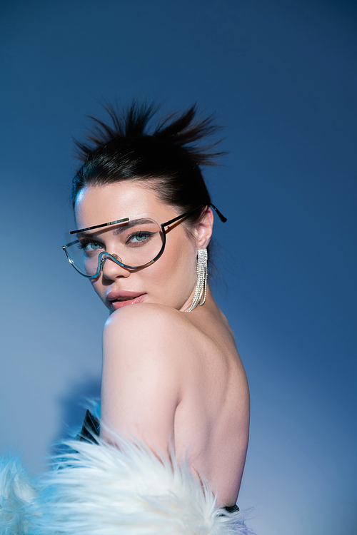 portrait of sensual woman with faux fur jacket and bare shoulder looking at camera in transparent sunglasses on blue background