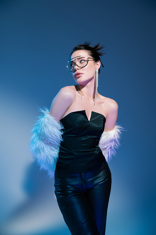 stylish woman in black corset and leather pants posing with white faux fur jacket on blue background
