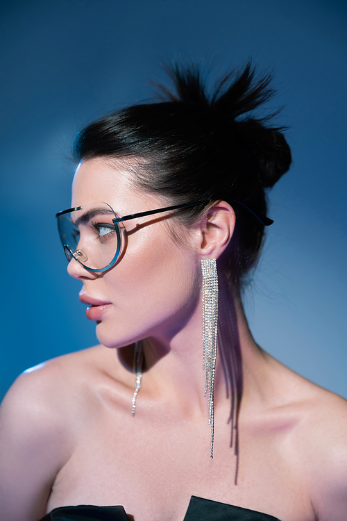 portrait of brunette woman in fashionable transparent eyeglasses looking away on blue background
