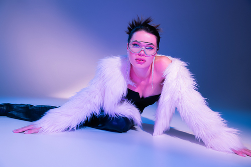 seductive woman in white faux fur jacket and transparent sunglasses posing on blue and purple background