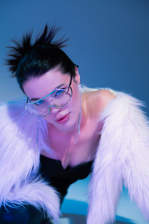fashionable woman in white faux fur jacket and transparent eyeglasses on blue background with purple light
