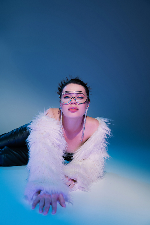 glamour woman in white faux fur jacket and transparent eyeglasses lying on blue background