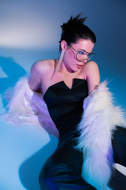brunette model in black corset and white faux fur jacket sitting on blue background