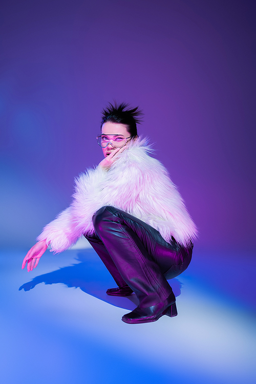 full length of brunette model in black leather pants and white furry jacket looking at camera on purple background