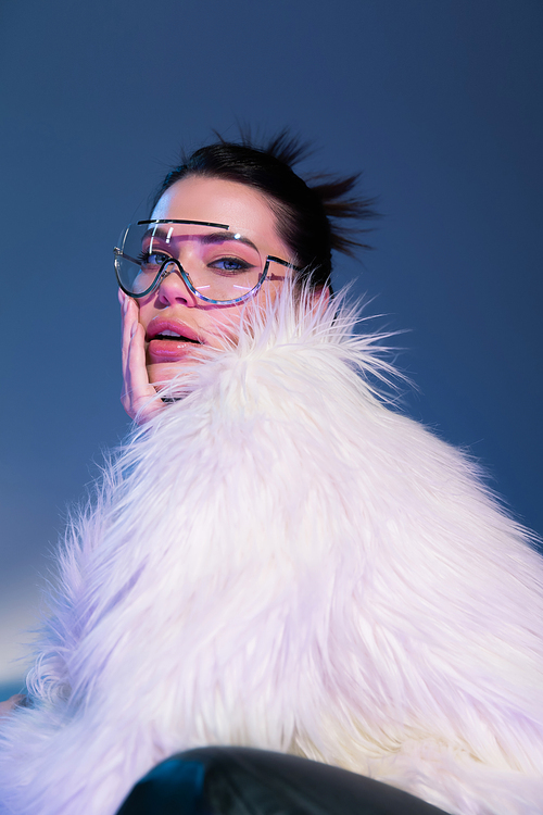 sensual brunette model in white faux fur jacket and transparent eyeglasses touching face on blue background