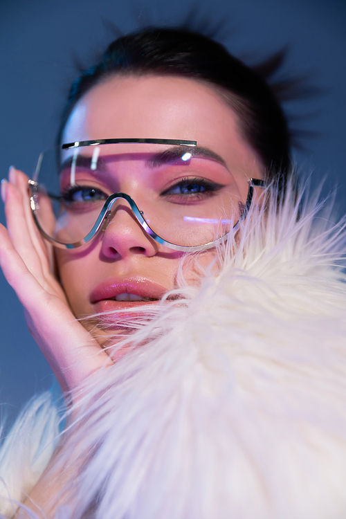 portrait of young woman in transparent sunglasses and white faux fur jacket looking at camera on blue background