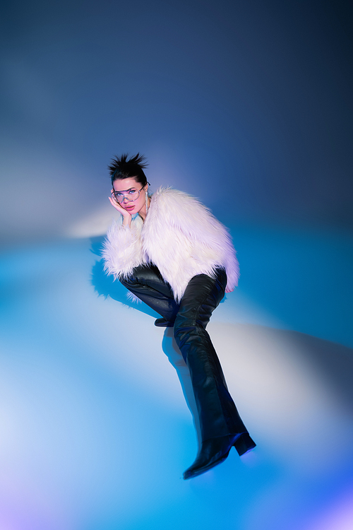 high angle view of fashionable woman in white furry jacket looking at camera while posing on blue background