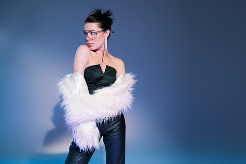 glamour model in black corset with leather pants looking away while posing in white faux fur jacket on blue background