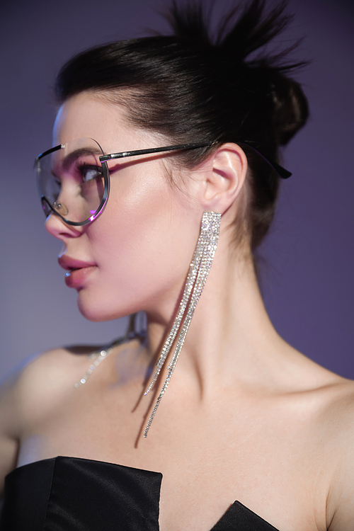 young brunette woman in shiny earrings and fashionable sunglasses looking away on purple background