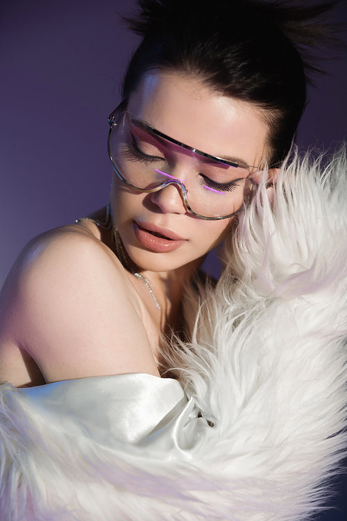 sensual brunette woman in trendy eyeglasses and white faux fur jacket posing on purple background