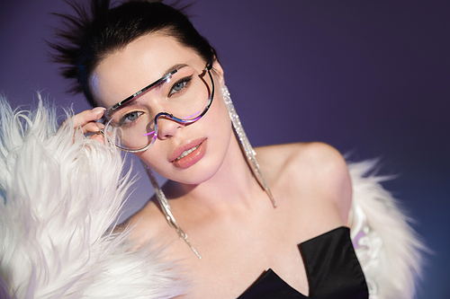 young woman in white faux fur jacket touching transparent eyeglasses on purple background
