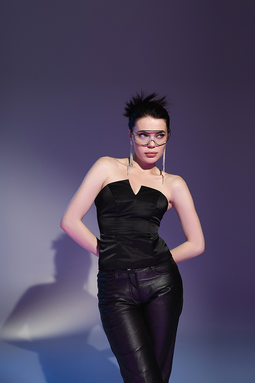brunette woman in fashionable eyeglasses and black corset posing with hands behind back and looking away on purple background