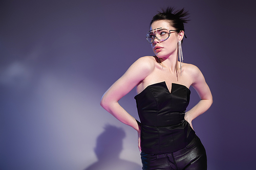 seductive woman in black corset and trendy sunglasses standing with hands on hips on purple background