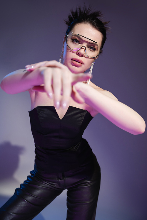 brunette woman in black corset and trendy sunglasses looking at camera and pointing with finger on purple background