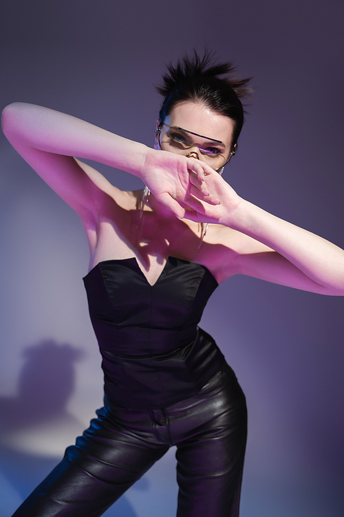 brunette woman in black corset and leather trousers obscuring face with hands on purple background