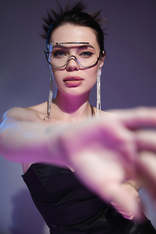 glamour woman in transparent sunglasses looking at camera near blurred outstretched hand on purple background