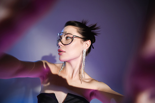 brunette woman in trendy sunglasses and shiny earrings looking away on blurred purple background