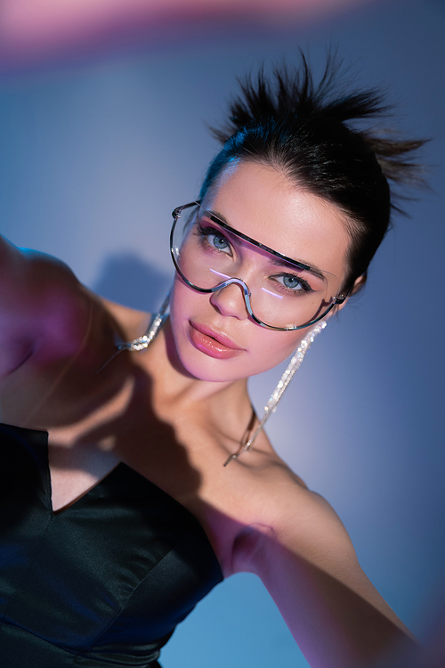 sensual woman in black corset and trendy sunglasses looking at camera on blue background