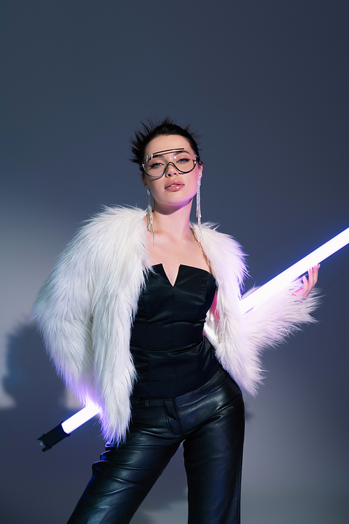 trendy woman in black corset and white faux fur jacket posing with fluorescent lamp on grey background