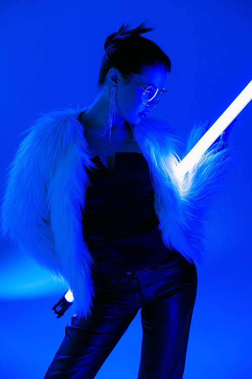 young woman in black corset and furry jacket posing with fluorescent lamp on bright blue background