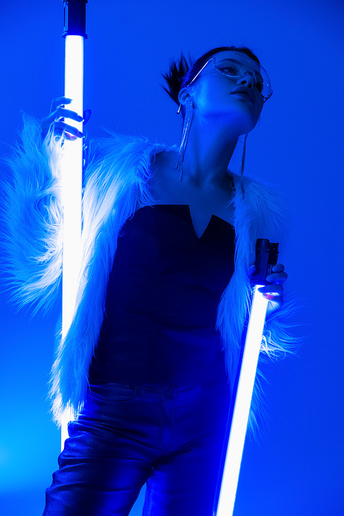 low angle view of trendy woman in faux fur jacket and black corset holding fluorescent lamps on blue background