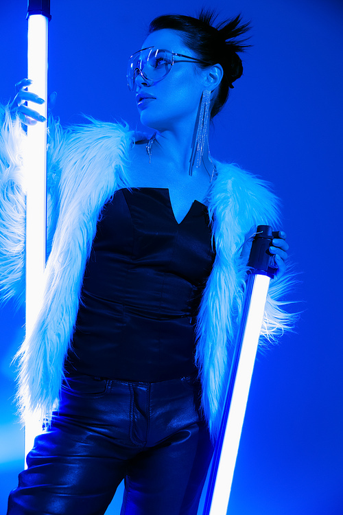 Low angle view of trendy woman in sunglasses and faux fur jacket holding lamps on blue background