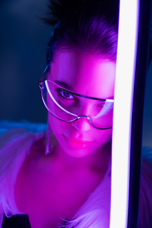 Trendy young woman in sunglasses looking at camera near lamp on navy blue background