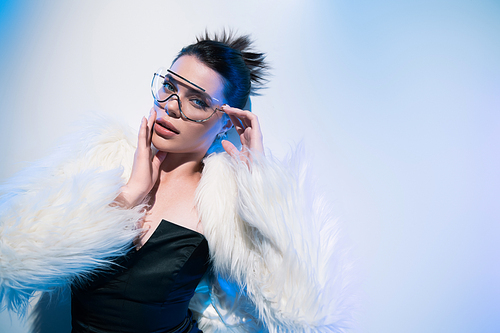 Top view of stylish woman in faux fur jacket and sunglasses looking at camera on white background with blue lighting