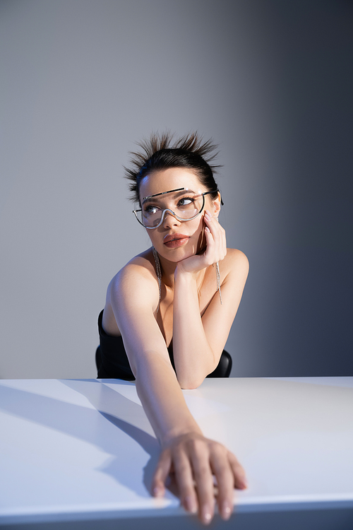 Trendy young woman in sunglasses looking away near table on grey background