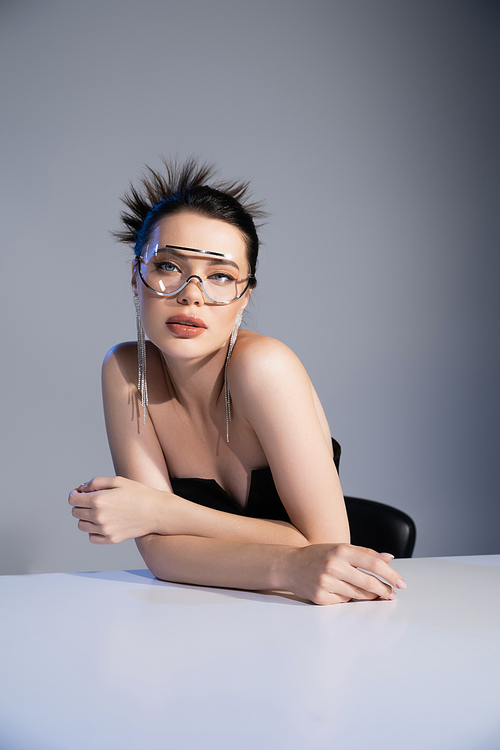 Stylish brunette woman in sunglasses looking at camera near table on grey background