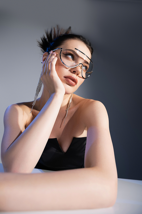 Trendy model in corset and sunglasses looking away near table on grey background