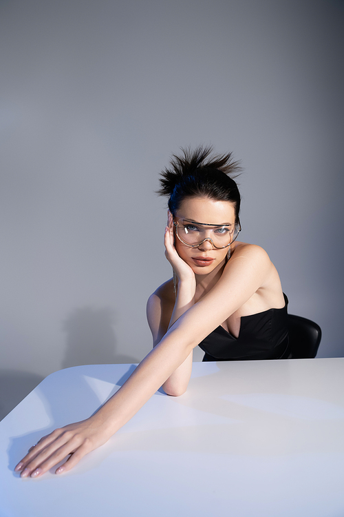 Pretty brunette woman in sunglasses touching face near table on grey background